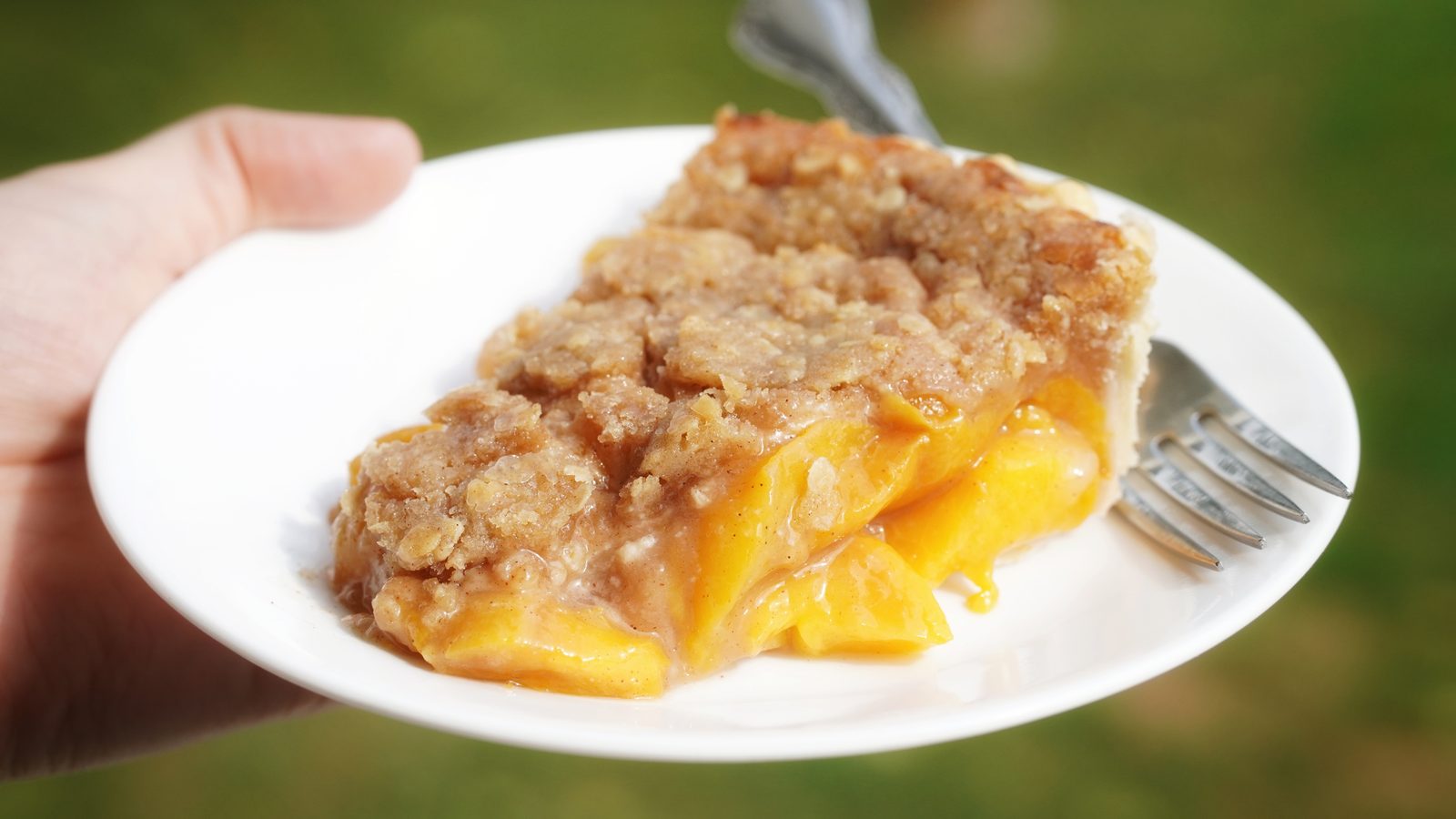 A slice of peach crumble pie on a white plate, held outdoors